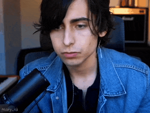 a young man wearing a denim jacket is sitting in front of a microphone and looking at the camera .