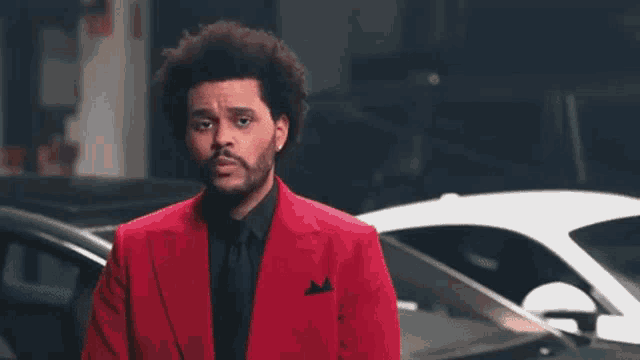 a man in a red suit and black tie stands in front of a white car .