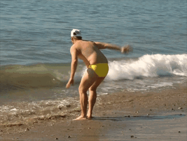 a shirtless man in yellow swim trunks and a white hat runs into the ocean