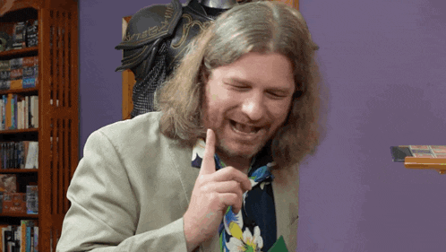 a man with long hair and a beard laughs in front of a purple wall