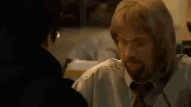 a man with blonde hair and a beard is sitting at a table with another man .