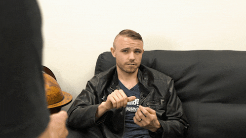 a man sitting on a couch wearing a black jacket and a blue shirt that says jigsaw on it