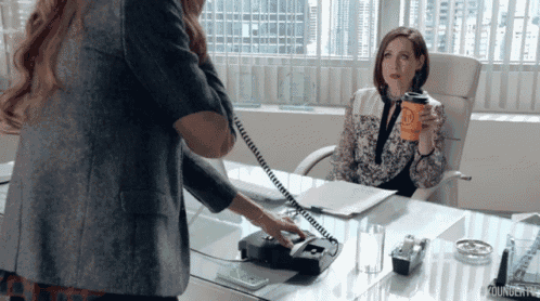 a woman talking on a phone while another woman sits at a desk with a cup of coffee