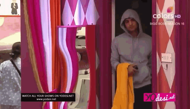 a man in a hooded sweatshirt is standing in a room with a sign that says colors hd bigg boss season 16