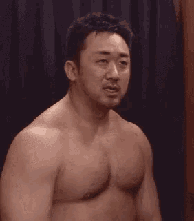 a shirtless man with a beard is standing in front of a black curtain and looking at the camera .