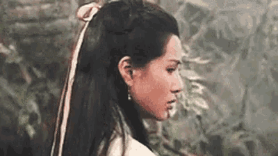 a woman with long black hair and a white ribbon in her hair is standing in a forest .