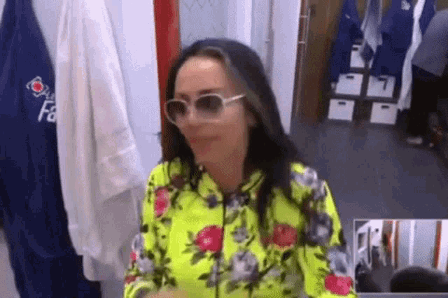 a woman wearing sunglasses and a yellow floral shirt is sitting in front of a mirror in a locker room .