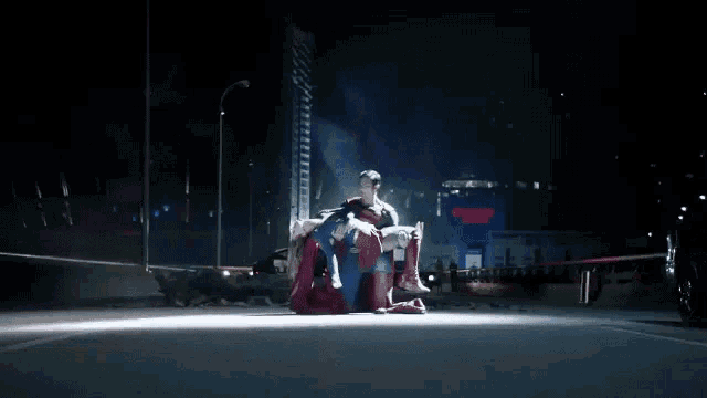 superman is holding a woman in his arms in a dark city .