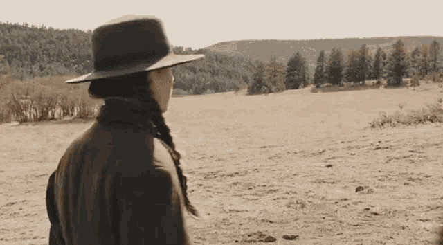 a woman in a hat stands in a field with mountains in the background