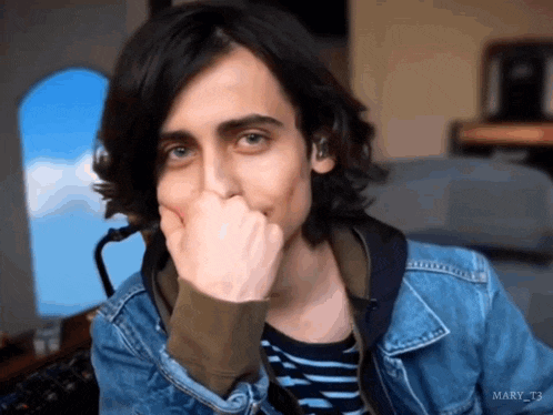 a young man with long hair is covering his mouth with his hand while wearing a denim jacket .