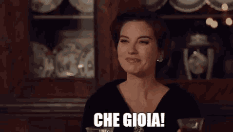 a woman in a black dress is sitting at a table with a glass of wine and says che gioia .