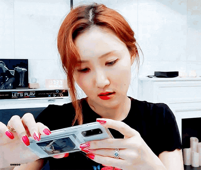 a woman with red nails is looking at her phone with a sign that says let 's play on the wall behind her