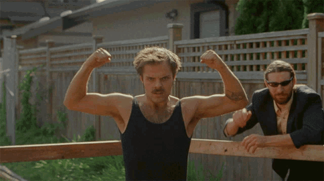 a man in a black tank top flexes his muscles while a man in a suit looks on