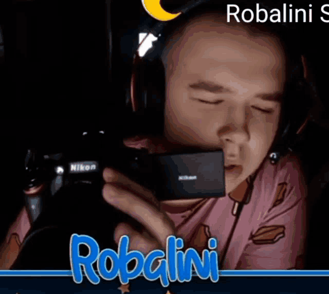 a man wearing headphones holds a nikon camera in front of a sign that says robalini