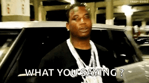 a man with a necklace around his neck is standing in front of a car and says `` what you saying ? ''