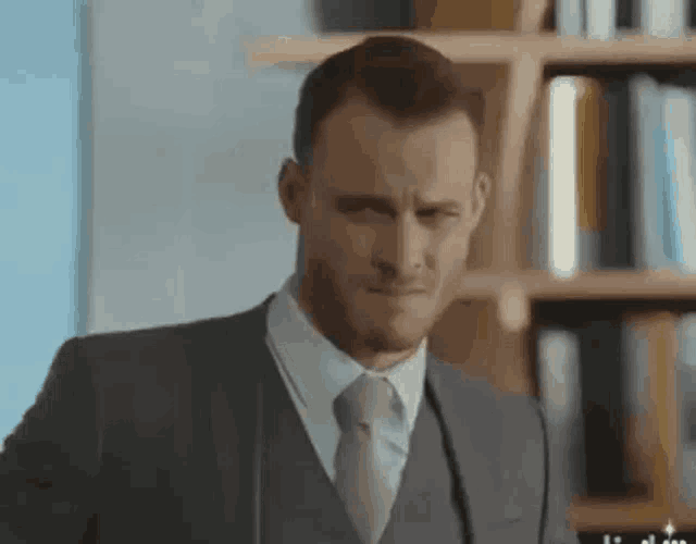 a man in a suit and tie is standing in front of a bookshelf and making a funny face .