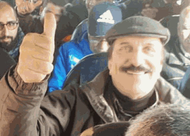 a man with a mustache is giving a thumbs up sign while sitting in a crowd of people .