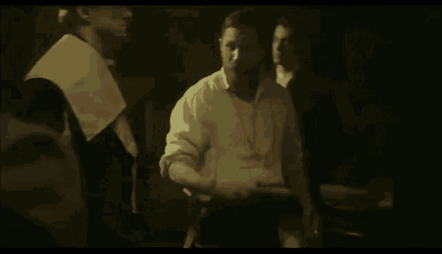 a man with a beard in a white shirt is standing in a dark room with other men .