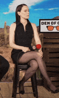 a woman is sitting on a stool with her legs crossed in front of a sign that says den of glasses