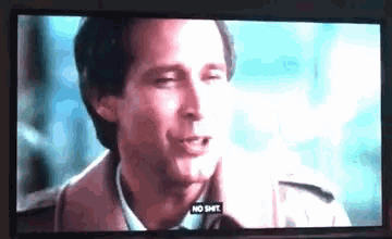a man in a trench coat is talking to another man on a television screen with a caption that says no shit .