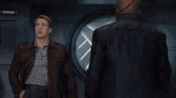 a man in a brown jacket stands in a room with a shield in the background