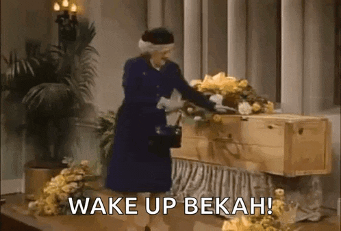 a woman in a blue dress is standing next to a wooden coffin with flowers on it .