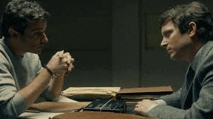 two men are sitting at a table with their hands folded in prayer .