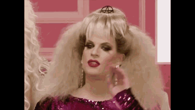 a drag queen wearing a pink dress and a wig is standing in front of a mirror .