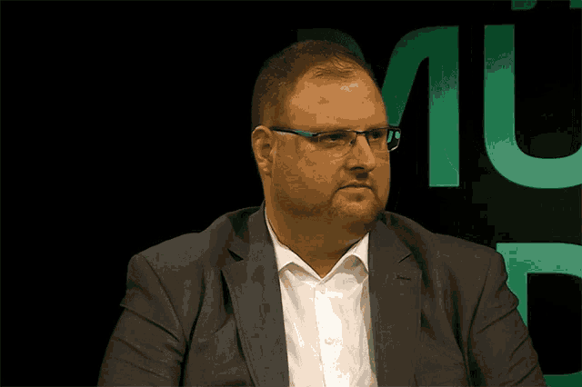 a man wearing glasses and a suit stands in front of a green m