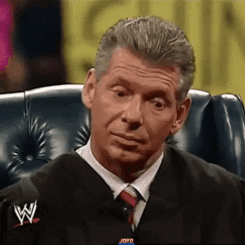 a man in a suit and tie is sitting in a chair with a wrestling logo on his jacket .