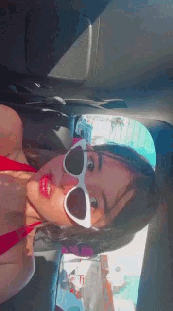 a woman in a red bikini and white sunglasses is taking a selfie in the back seat of a car .