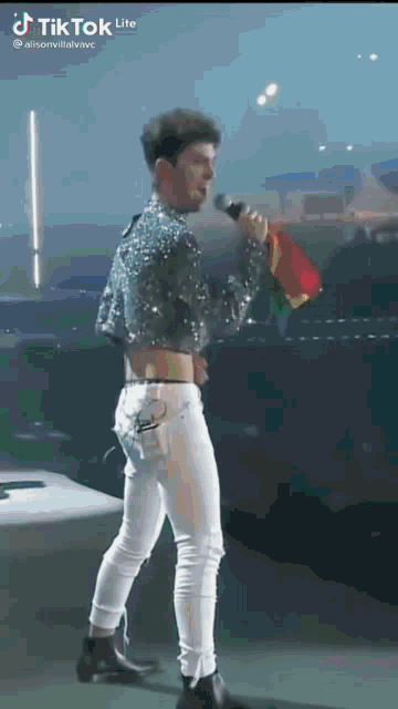 a man is singing into a microphone on a stage while wearing a crop top and white jeans .