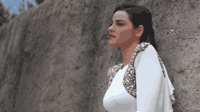 a woman in a white dress with gold beads on the shoulder is leaning against a wall