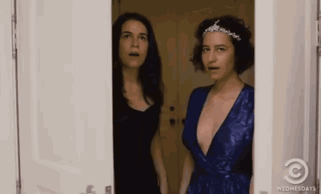 two women are standing next to each other in a doorway . one of the women is wearing a blue dress .