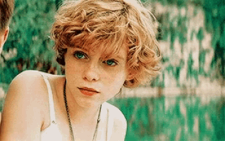 a young girl with curly hair and green eyes is wearing a white tank top .