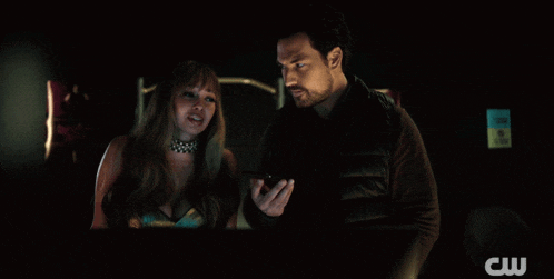 a man and a woman are looking at a cell phone with the cw logo in the corner