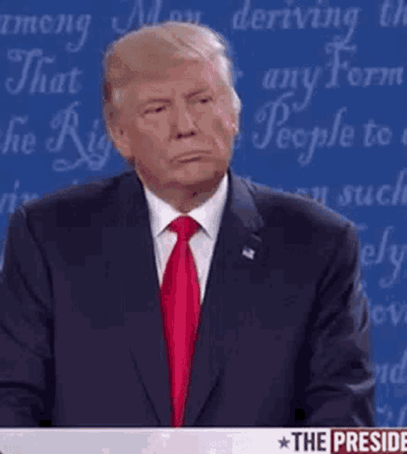 donald trump is wearing a suit and tie and making a funny face while standing in front of a blue wall .