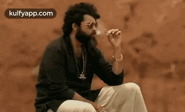 a man with a beard and sunglasses is sitting on the ground smoking a cigarette .