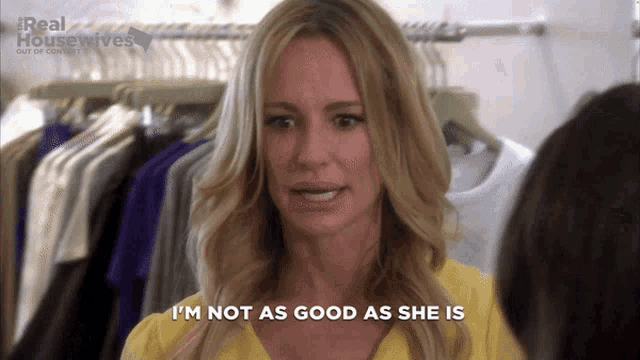 a woman says " i 'm not as good as she is " in front of a rack of clothes