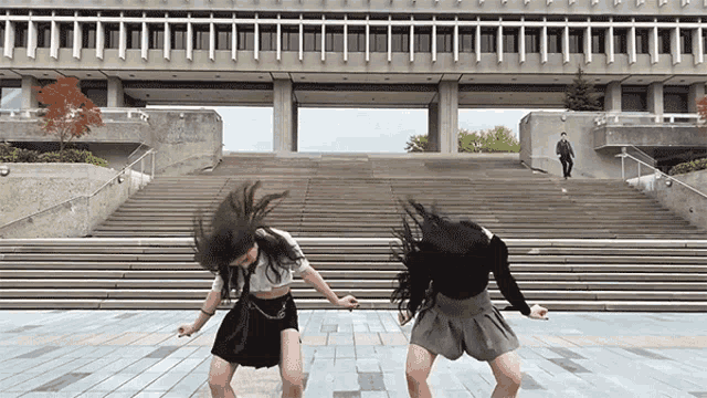 two girls are dancing in front of a building with stairs and a man is walking down them