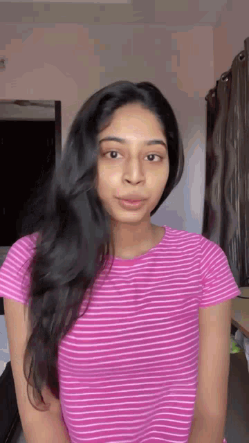 a woman with long black hair is wearing a pink striped shirt and making a funny face .