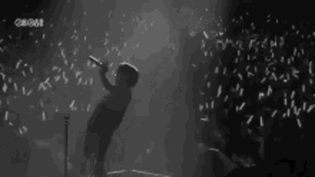a black and white photo of a man standing on a stage holding a microphone in front of a crowd .