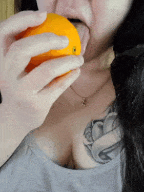 a woman with a tattoo on her chest is biting an orange