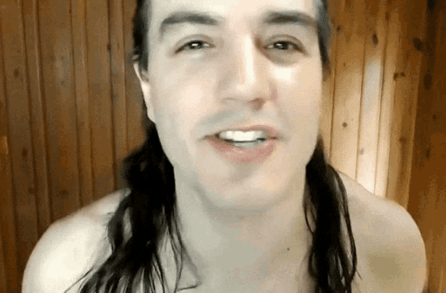 a man with a ponytail is smiling in front of a wooden wall