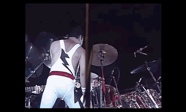 a man in a white tank top with a lightning bolt on it is standing on a stage playing drums .