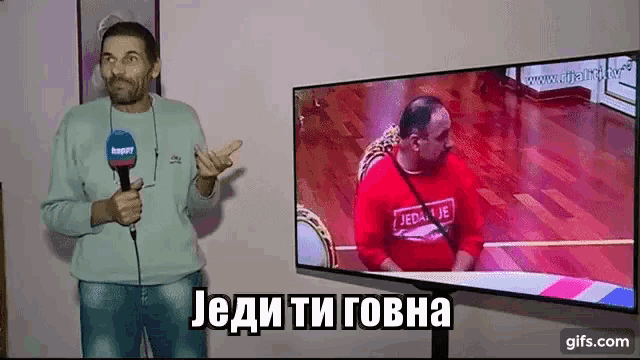 a man is holding a microphone in front of a tv screen with a man in a red shirt on it .