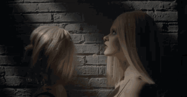 two blonde women looking at each other in front of a white brick wall