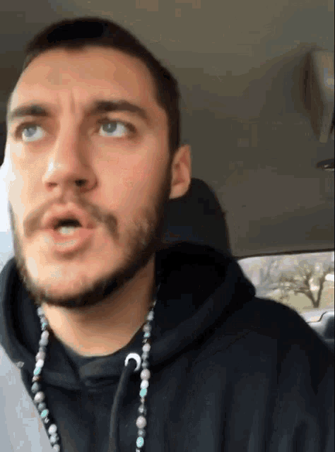 a man with a beard is wearing a black hoodie and a necklace with the letter c on it