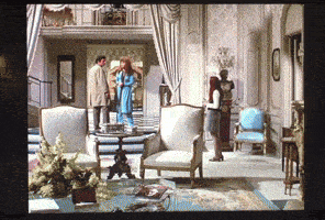 a woman in a blue dress stands in a living room talking to a man