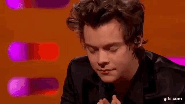 harry styles is wearing a black jacket and smiling while sitting in front of a colorful background .